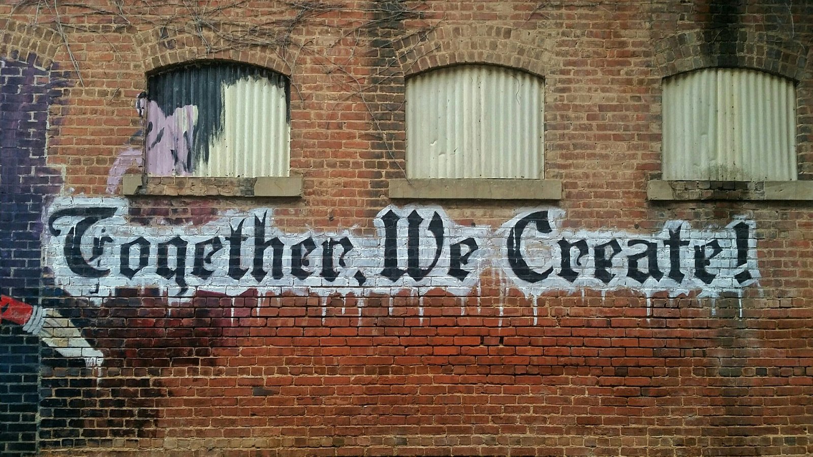 "Together, We Create!" along with a dripping paint brush, painted on a red brick wall.